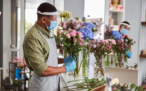 Junger Blumenerkäufer steht mit Maske und Visier und bindet in seinem Laden einem Blumenstrauß