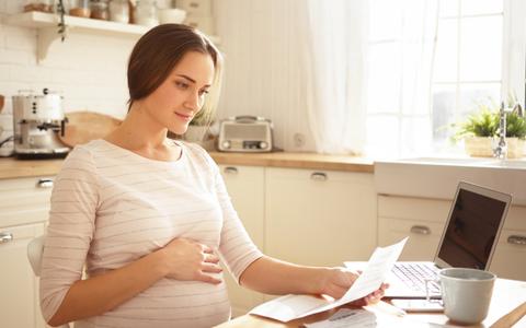 schwangere Frau sitzt am Küchentisch mit Laptop und bearbeitet Rechnungen