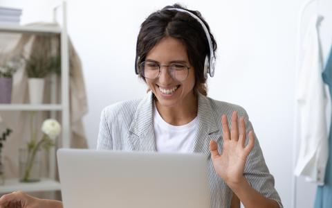 Titelbild: Junge Frau mit Kopfhörer im Homeoffice winkt vor Laptop bei einer Videokonferenz
